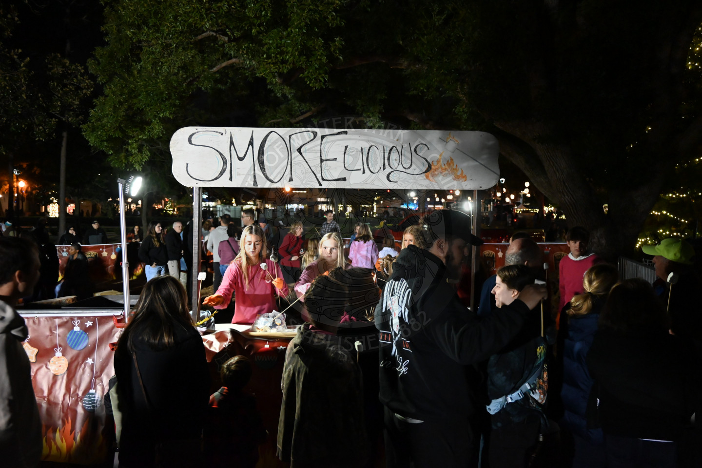 A stand with people handing out utenstils to make smores. there is a sign above the stand that says "smorelicious".