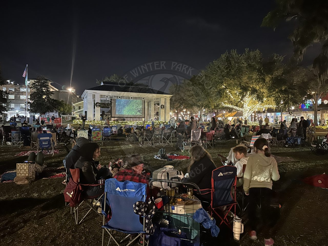 groups of people sitting in chairs or on picnic blankets in the park watching a movie on a big screen.