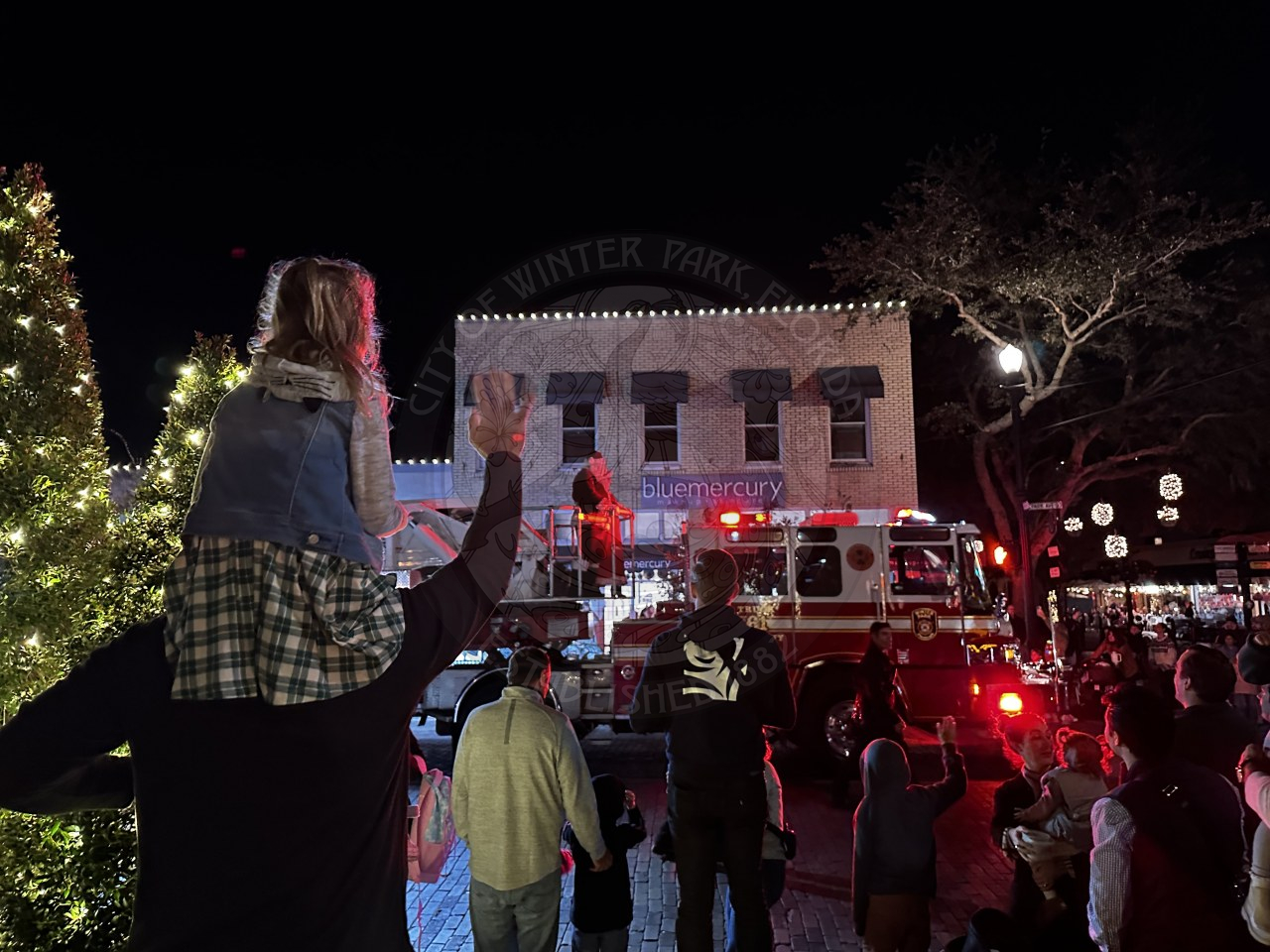 a man holding a little girl on his shoulders and waving to Santa Clause who is passing by on a firetruck.