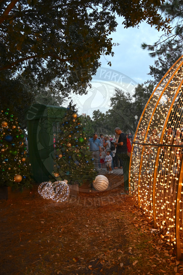 Lighted up arch way with christmas trees next to it and people walking around behind both of them.