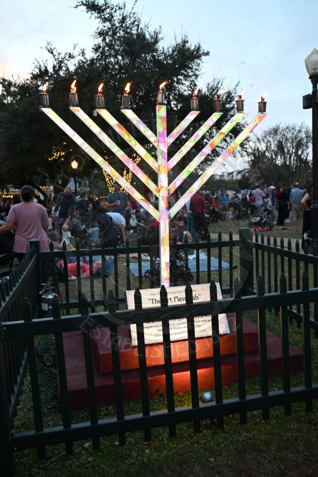 A colorful lit menorah in the middle of a grass area with a fence surrounding it.
