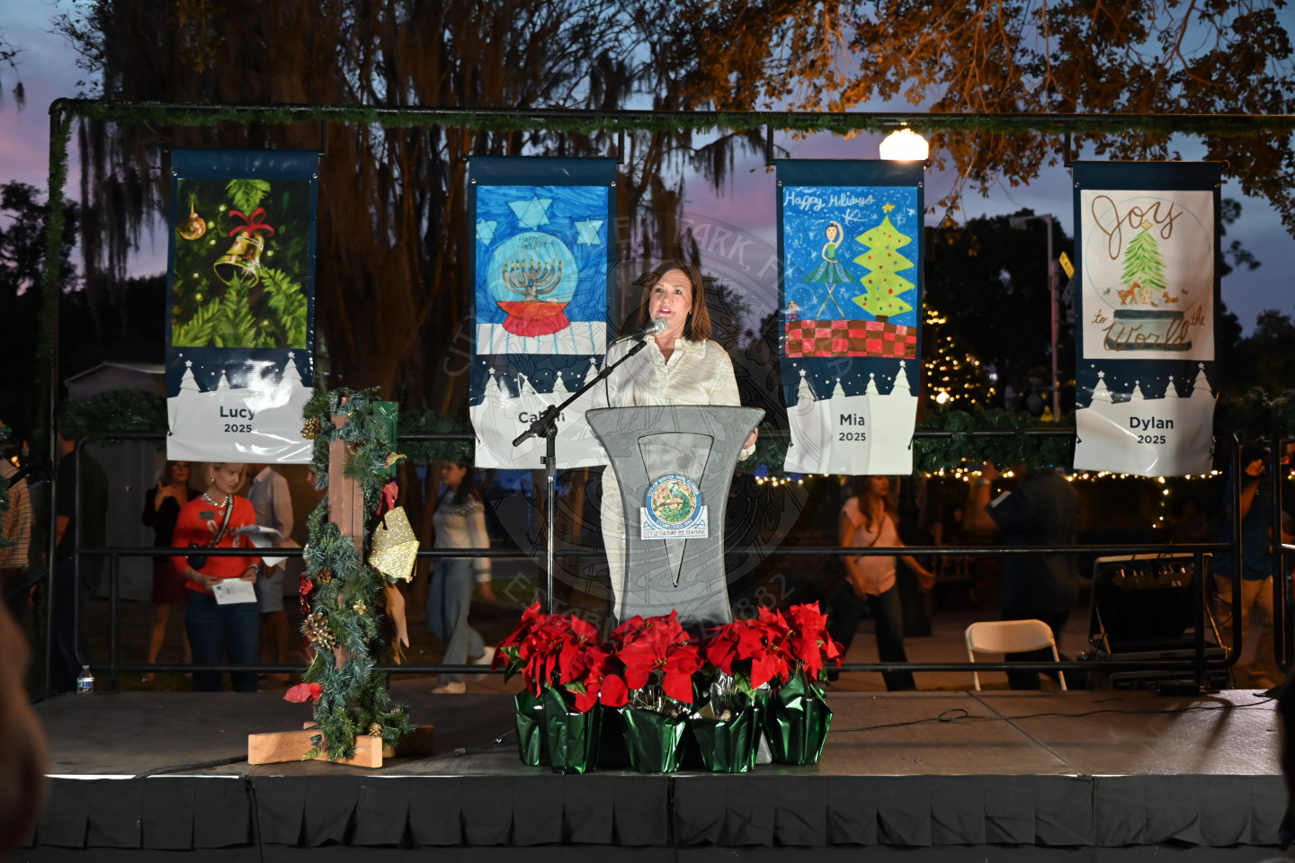 a woman standing on a stage behind a podium talking into a microphone with drawings children drew hanging up behind her.