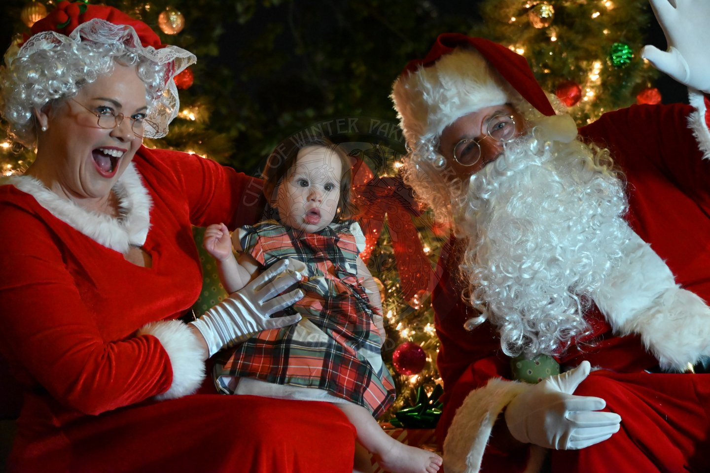 Mrs. Claus holding a baby in her lap with Santa Claus next to her smiling.
