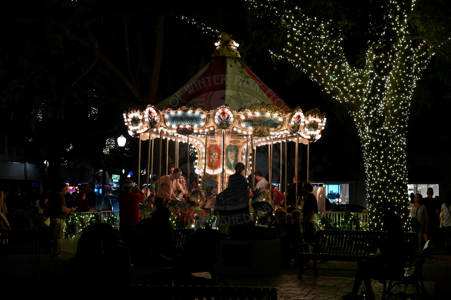 at night, a lit up carousel with children and parents on the ride. There is a wrapped up tree with lights on it next to the carousel.