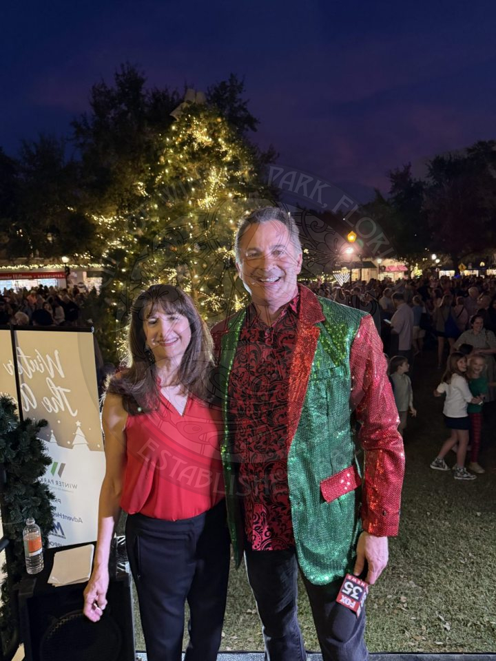 A man and woman standing next to each other in front of a Christmas tree smiling.