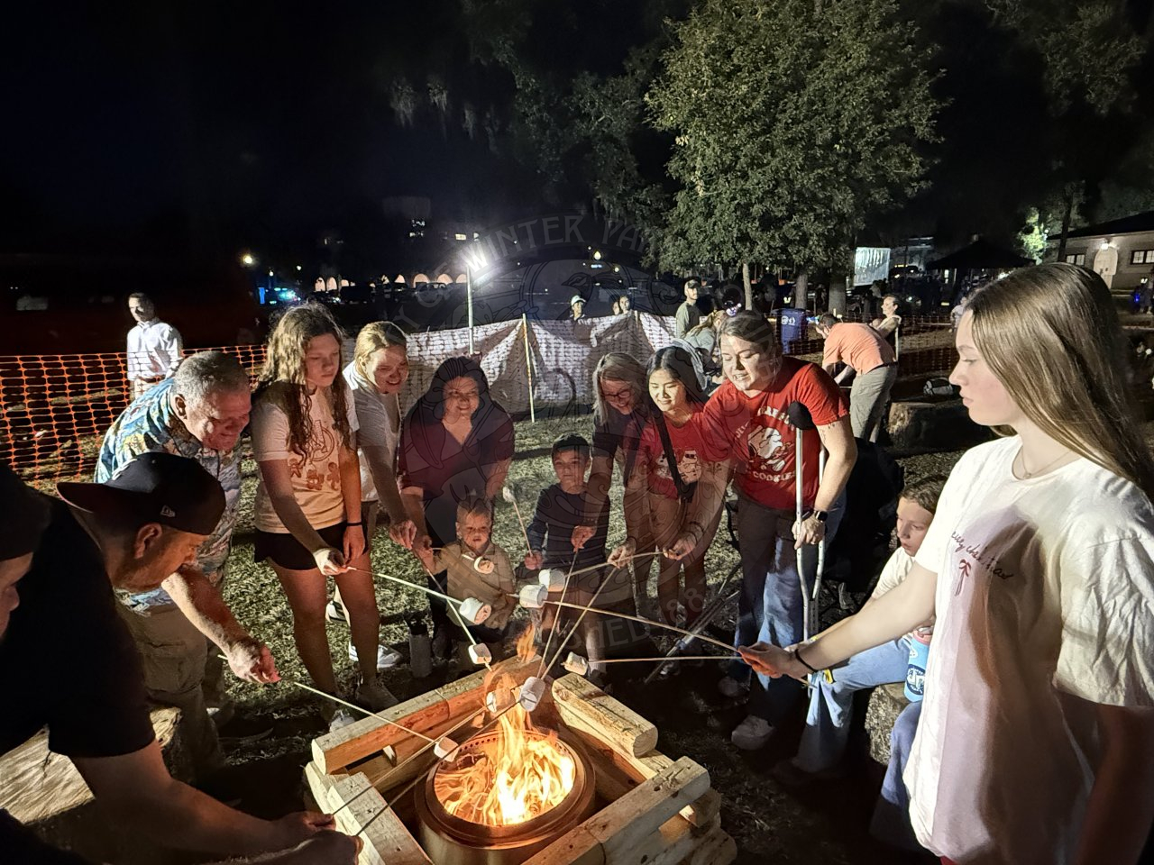a group of adults and children standing around a campfire holding sticks with marshmallows on the end of them roasting over the fire.