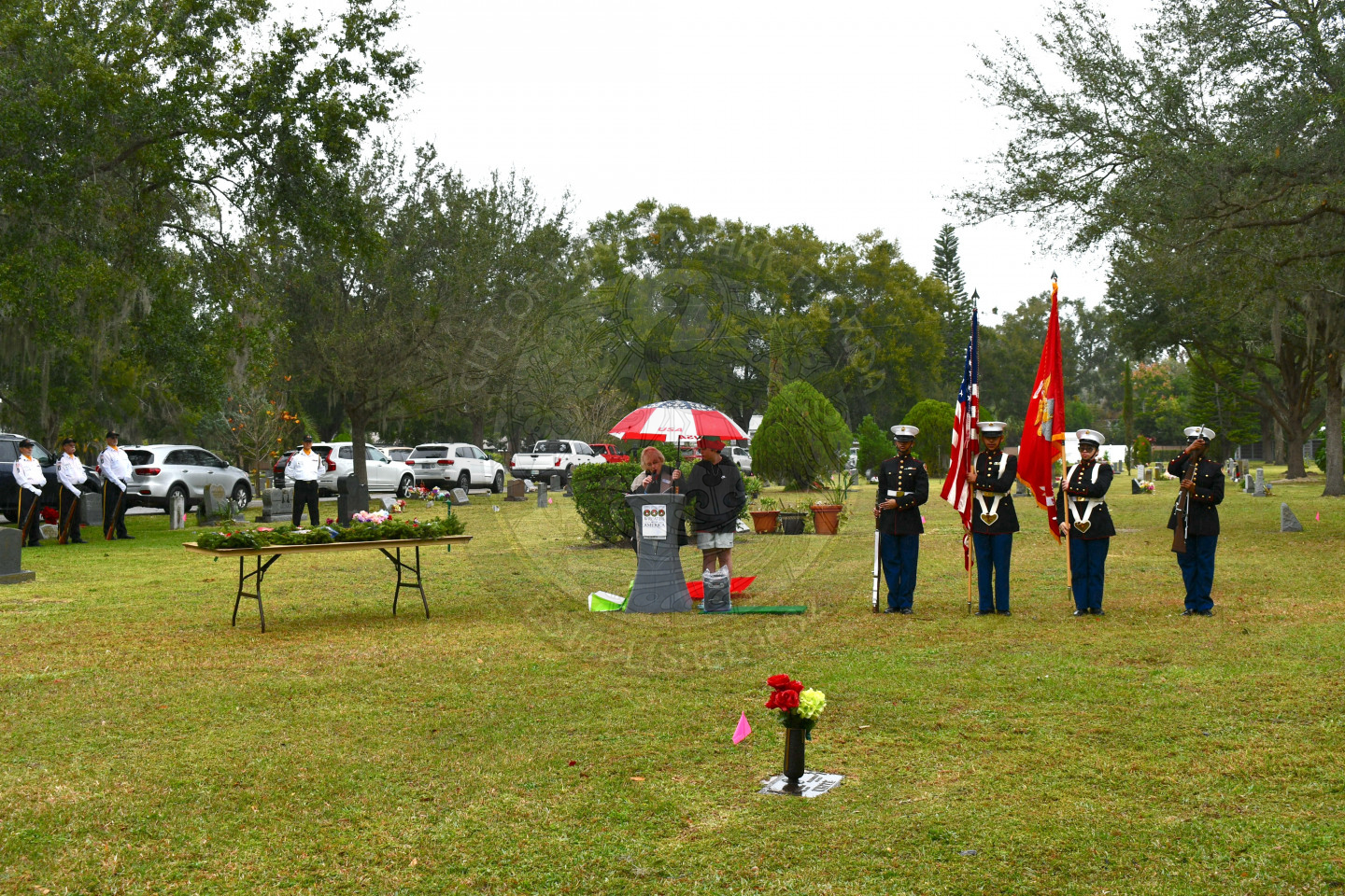 At a cemetary. on the left hand side is a 6 foot table with wreaths on it, in the middle is a podium with a woman speaking into a microphone and a boy standing next to her holding an umbrella for her, to the right is the color guard.