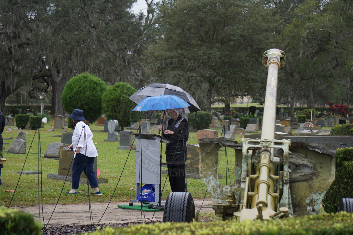 A woman standing behind a podium holding an umbrella to protect her from the rain while she speaks to a crowd of people.