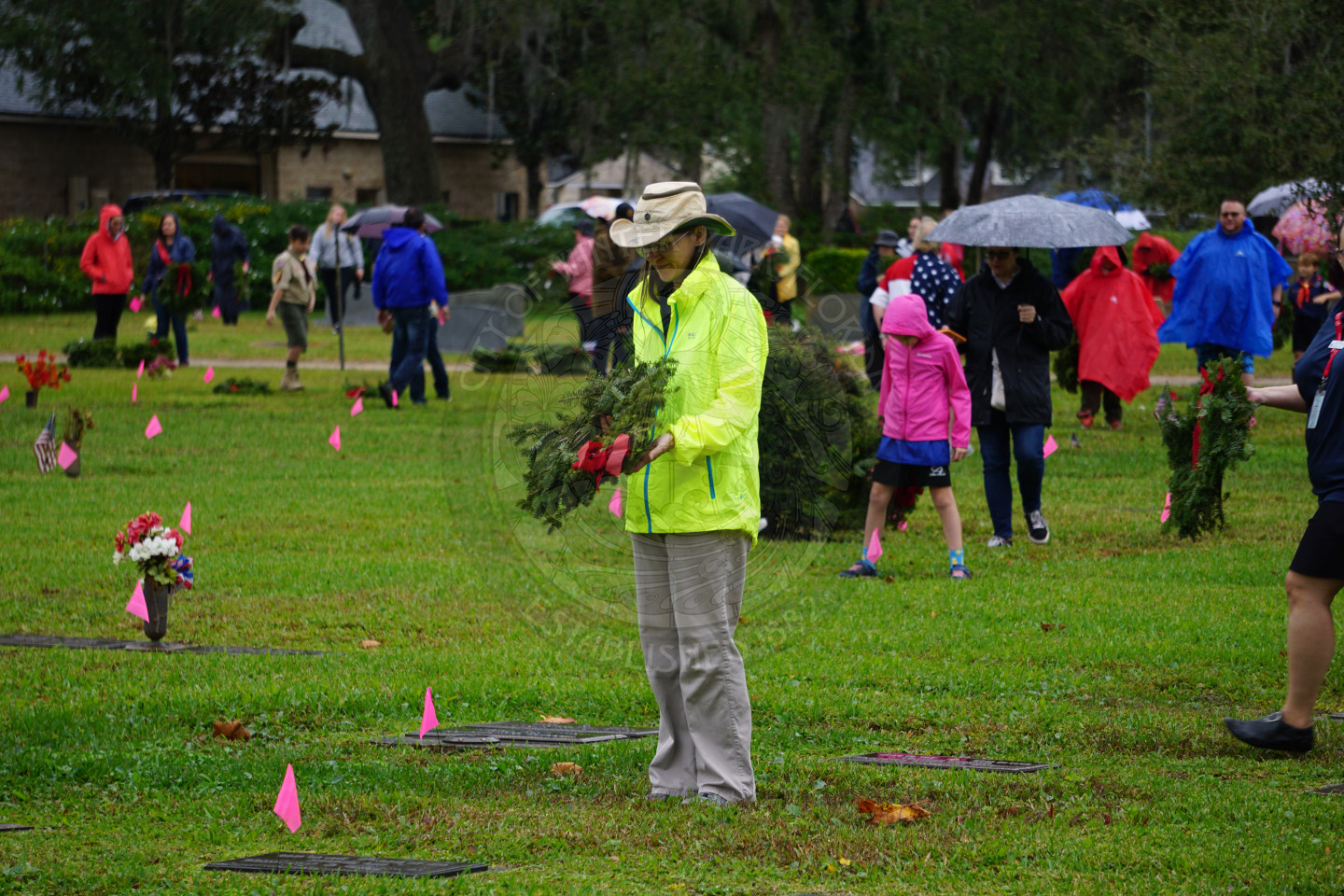 A woman standing over a grave as she is about to bend down to place a wreath on a grave.