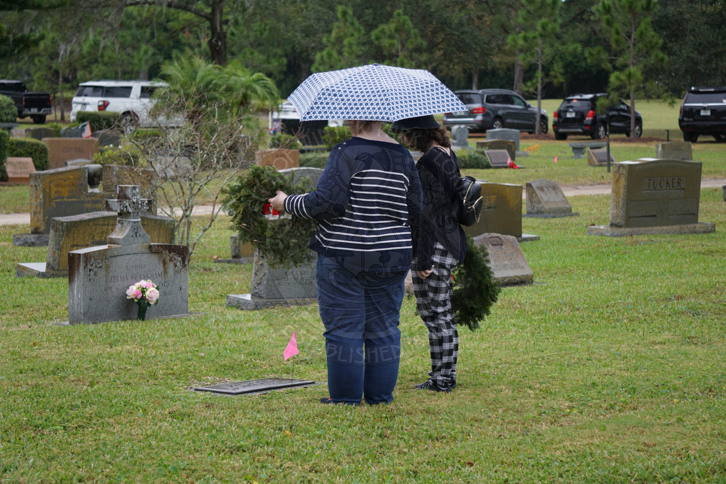 two women standing next to each other. One is holding an umbrella and the other is holding a wreath as they look down at a grave.