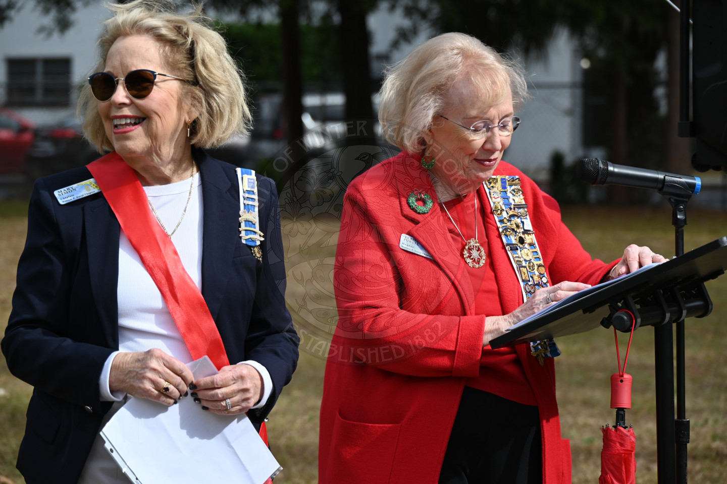 an older lady talking into a microphone looking through papers that are on a stand in front of her. There is another older lady standing next to her holding a peice of paper.