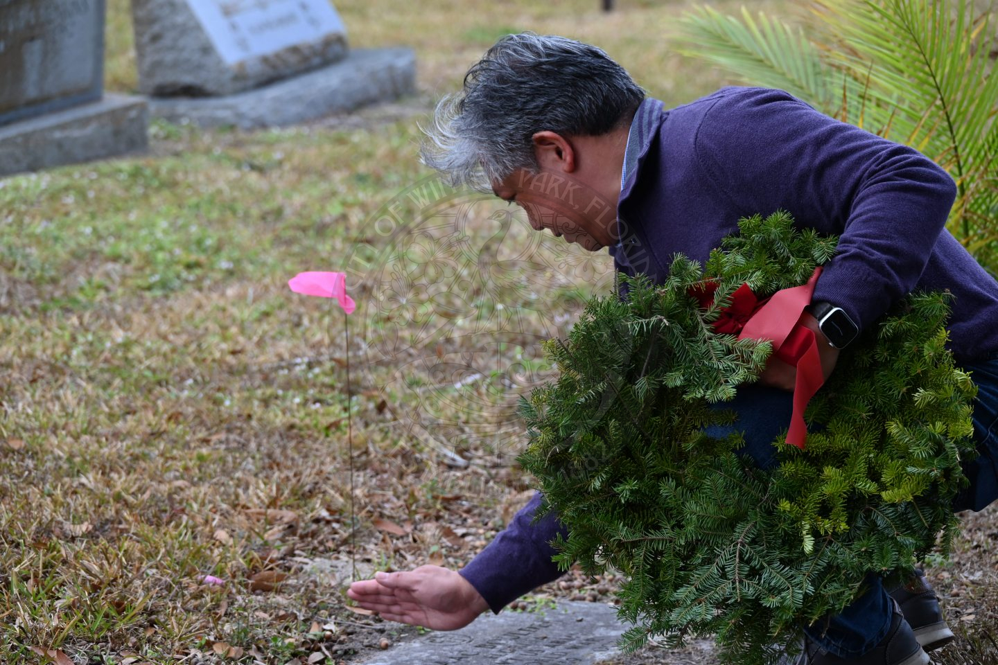 a man squatting down placing a wreath on a grave but also wiping off the dirt on the grave.
