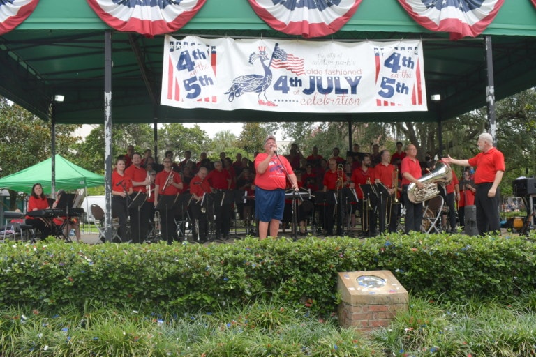 Brass band and choir members on park stage with emcee and conductor for 4th of July event