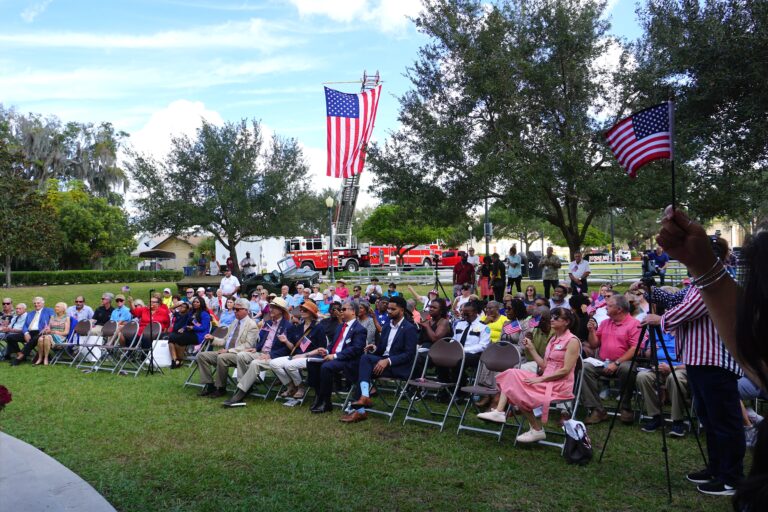 people sitting in chairs that are in multiple rows in green grass. There is a big american flag hanging from a firetruck swaying in the wind.