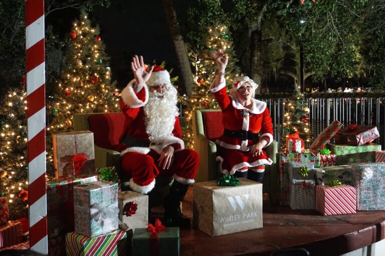 Santa and Mrs. Claus sitting in red chairs on a stage with a bunch of presents surrounding them waving to the crowd.