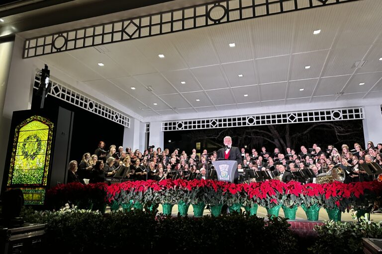 the director of Bach standing on the stage behind a podium with the choir behind him waiting to perform. To the left of the stage is a yellow/green stained glass window with a wreath on it.
