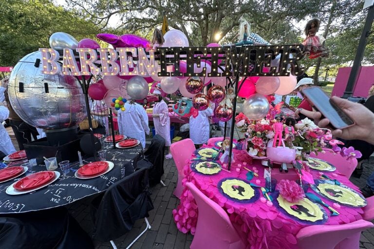 two round tables pushed together to make Barbenheimer which is the movies Barbie and Oppenhiemer put together. One table has scientific equations and a giant metalic globe in the middle and the other table is all pink with barbie dolls and barbie plates on it.