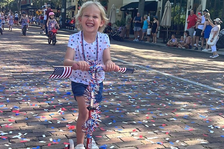 a little girl wearing red, white and blue riding her scooter that is decorated with American garland.