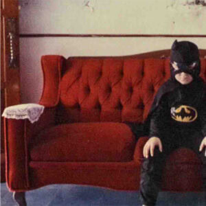 a little boy dressed in the batman costume sitting on a red couch with his hands placed on his knees.