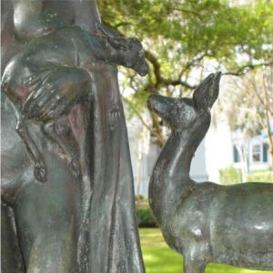 a lady holding a baby deer in her left arm with the mother deer looking at the baby deer.