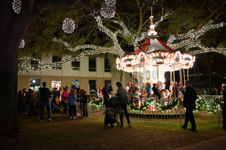 a carousel with a a tree with lights on the branches behind it. People are waiting in line to get on the carousel with kids on it.