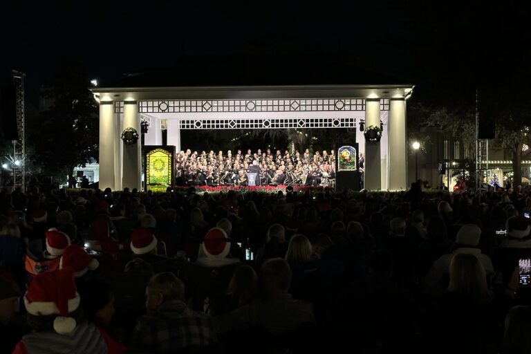 An outside stage with two tiffany stained glass windows on the left and right side of the stage. In the middle is a symphonic band sitting and playing their instruments to perform a concert.