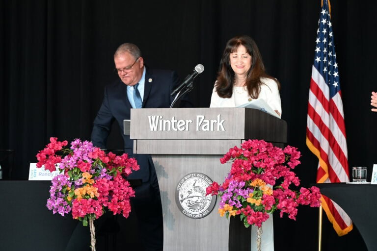 a woman standing behind a podium with two flower pots next to the podium smiling while looking down at the podium. There is also an American flag behind the woman.