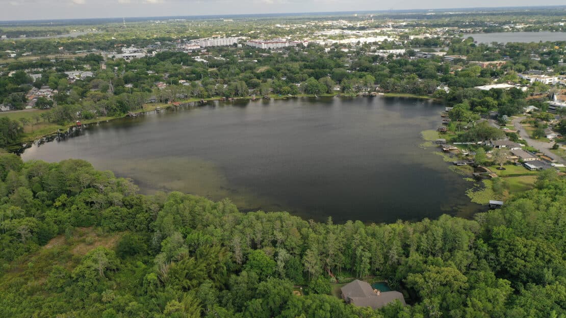 Aerial view of a circular lake, part of the region's beautiful lakes, surrounded by green trees and houses, with a cityscape in the background under a partly cloudy sky.