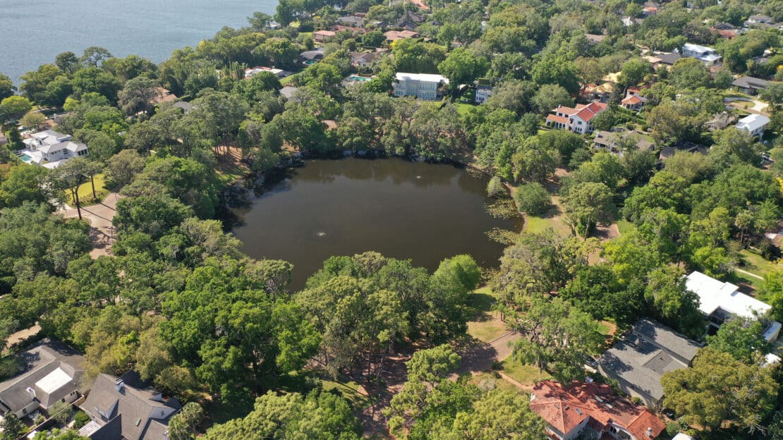 Aerial view of a small, round pond surrounded by dense green trees and residential houses, with a larger body of water—one of the local lakes—visible in the top left corner, providing general information about the area’s landscape.