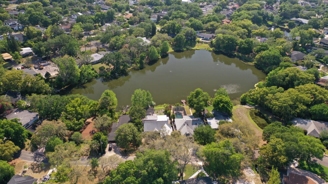 Aerial view of a suburban neighborhood surrounding a small, tree-lined lake, with houses, roads, and lush green foliage throughout the area—offering general information about tranquil lakeside living.