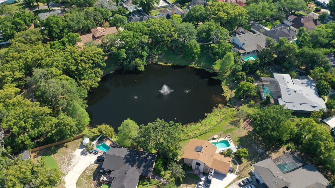 Aerial view of a residential neighborhood with houses surrounding a small lake featuring a fountain in the center, lots of trees, and some houses with pools in their backyards—perfect for those seeking general information about scenic Lakes communities.