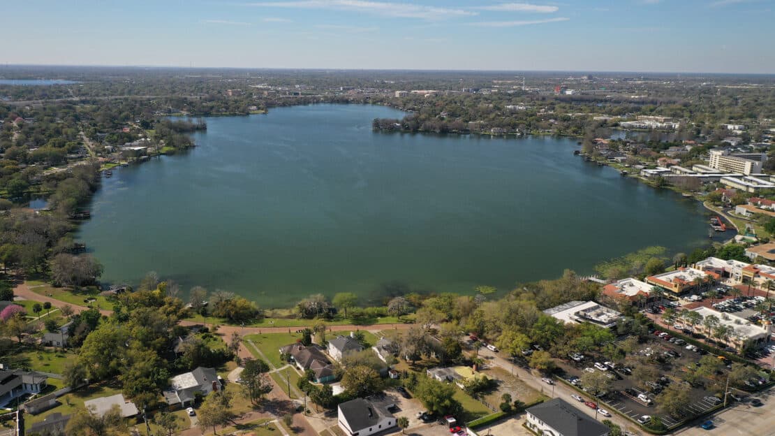 Aerial view of a large circular lake—part of the city’s scenic lakes—surrounded by houses, trees, and buildings on a sunny day, with a sprawling cityscape in the background.