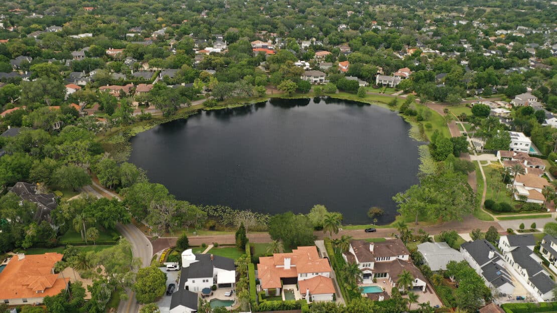 Aerial view of a suburban neighborhood with a circular lake at the center, surrounded by houses, trees, and greenery. The rooftops of the homes are visible, with roads curving around the lake, offering general information about residential lakes.