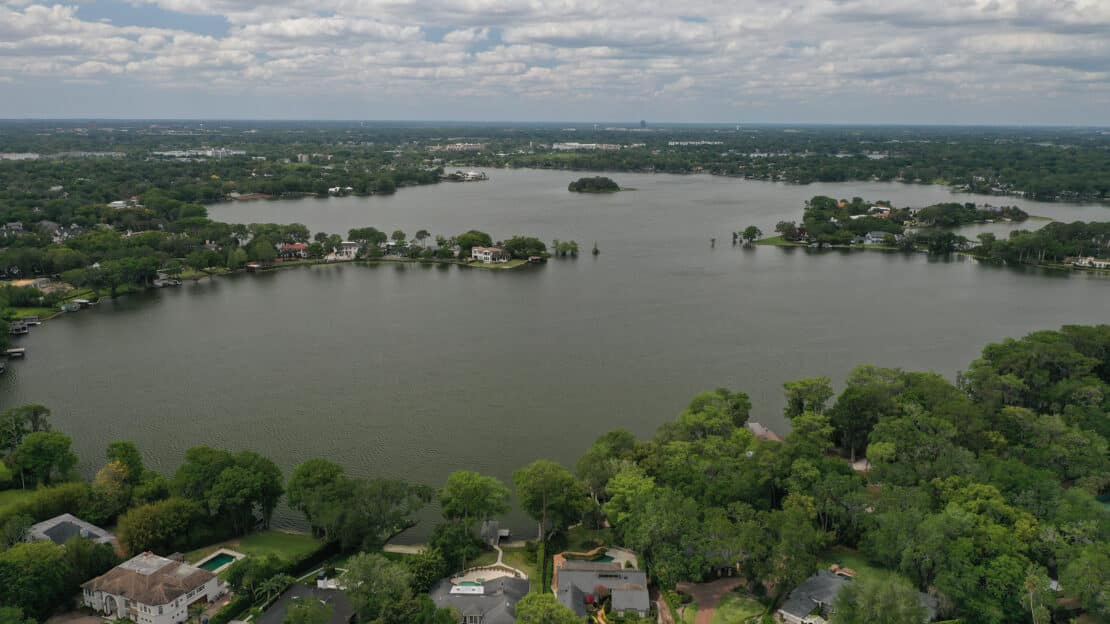 Aerial view of a large lake, offering general information on lakes, surrounded by green trees and houses with a small island in the center. The sky is partly cloudy and the horizon stretches into the distance.