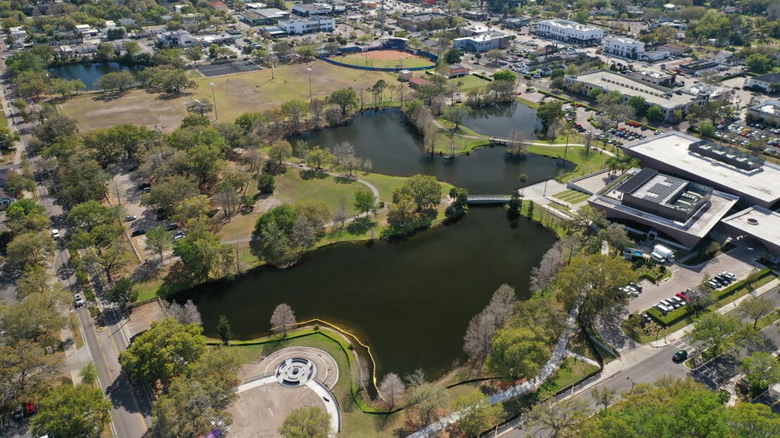 Aerial view of a city park with lakes, green spaces, winding paths, a circular plaza, and nearby buildings and parking lots surrounded by trees and roads offers general information about the park’s layout.