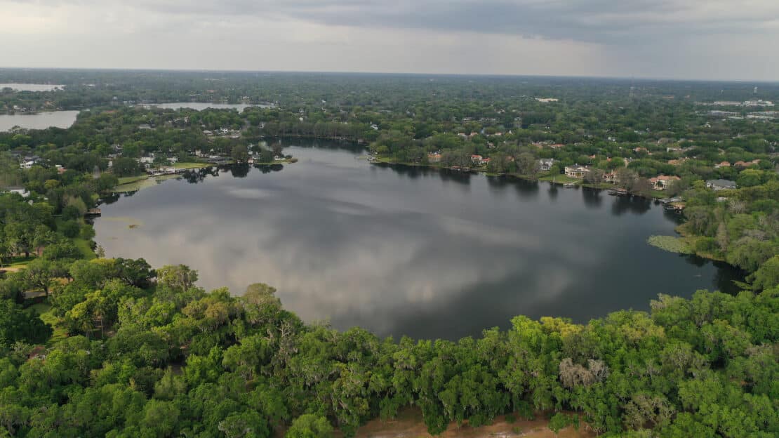 Aerial view of a large, tree-lined lake surrounded by residential houses, with dense green foliage in the foreground and a cloudy sky reflected on the water's surface—ideal for those seeking general information about scenic lakes.