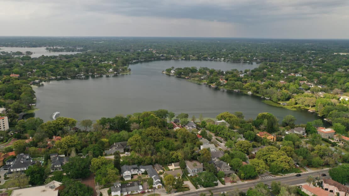 Aerial view of a residential neighborhood with many trees surrounding expansive lakes on a cloudy day, houses scattered along the shoreline and leafy streets visible in the foreground, offering general information about the area’s natural beauty.