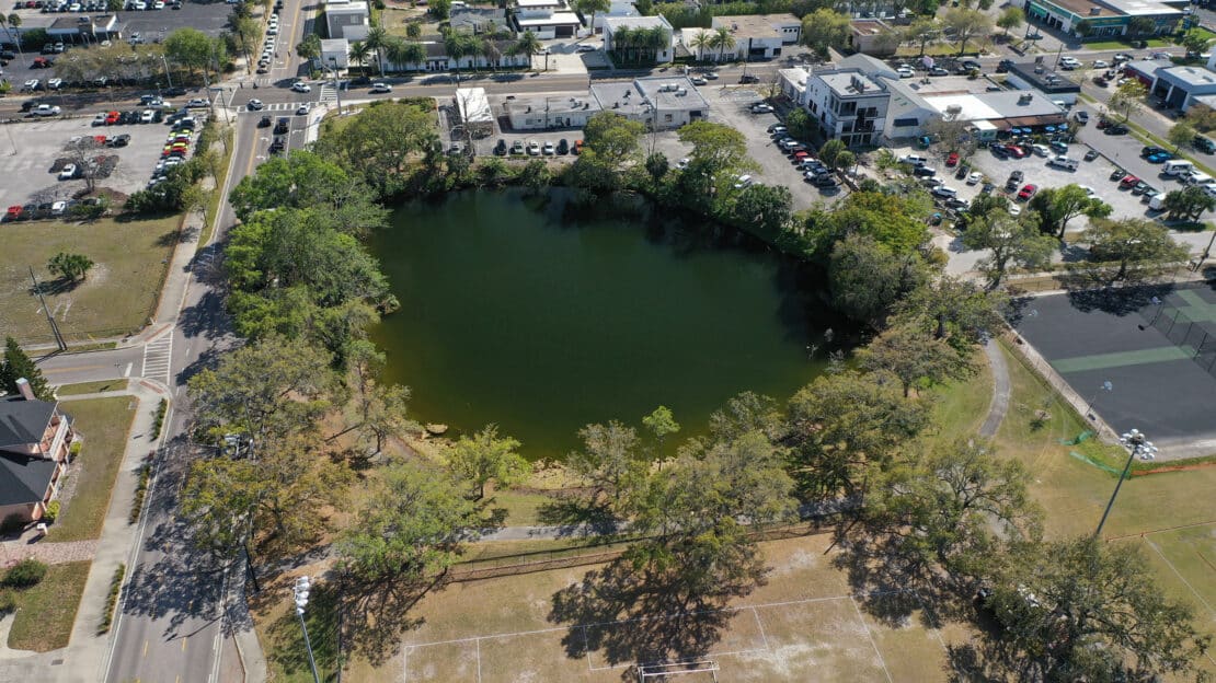 Aerial view of a circular pond, similar to small lakes, surrounded by trees with nearby buildings, parked cars, and a sports court. The area offers general information about urban or suburban neighborhood layouts.