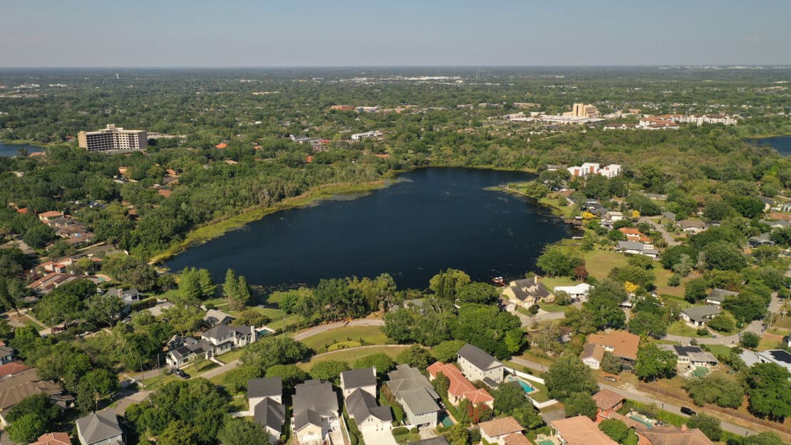 Aerial view of a small lake surrounded by trees and residential houses, with lakes dotted throughout the suburban cityscape and distant buildings visible under a clear blue sky, offering general information about the area’s landscape.
