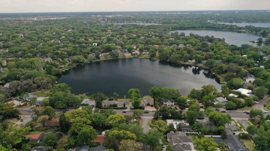 Aerial view of circular lakes surrounded by houses and dense green trees in a suburban neighborhood, with more lakes and greenery in the distant background under a cloudy sky, offering general information about local landscapes.