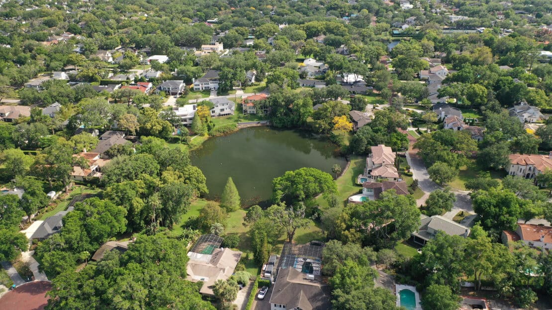 Aerial view of a residential neighborhood with tree-lined streets and houses surrounding a small pond, offering general information on the area's dense greenery, scattered swimming pools, and nearby lakes.