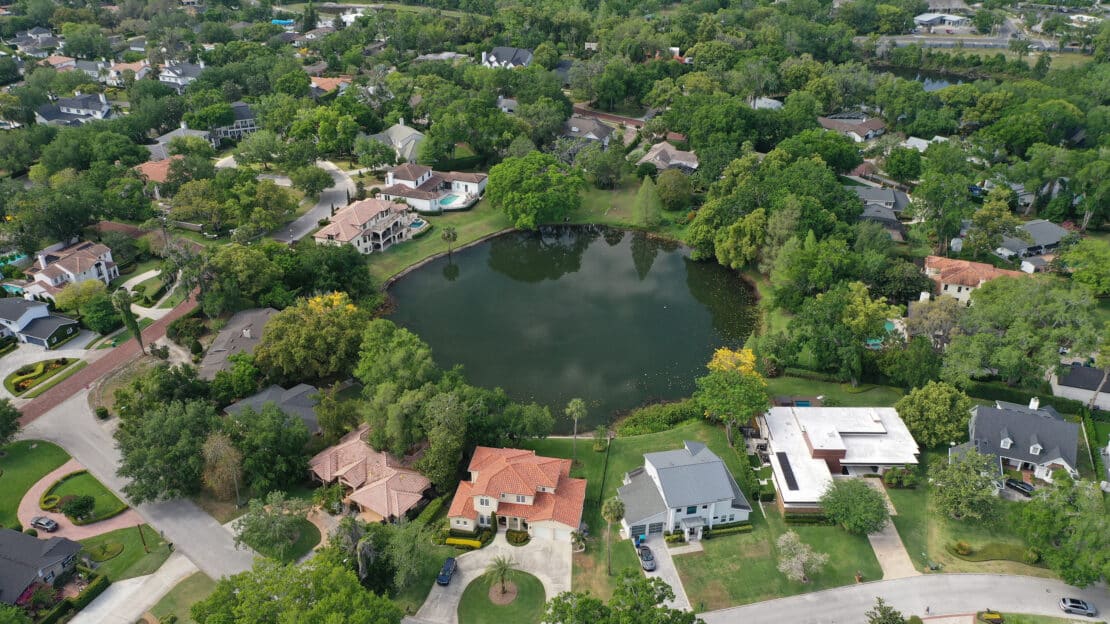 Aerial view of a suburban neighborhood with houses surrounding a small, tree-lined lake. The curved streets and lakes create a peaceful setting, enhanced by lush greenery and well-maintained lawns.