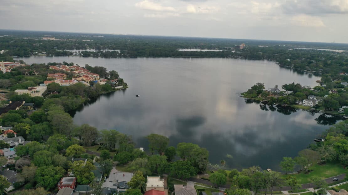 Aerial view of a large lake surrounded by residential neighborhoods with houses, trees, and greenery under a cloudy sky, offering general information about how lakes enhance local landscapes.