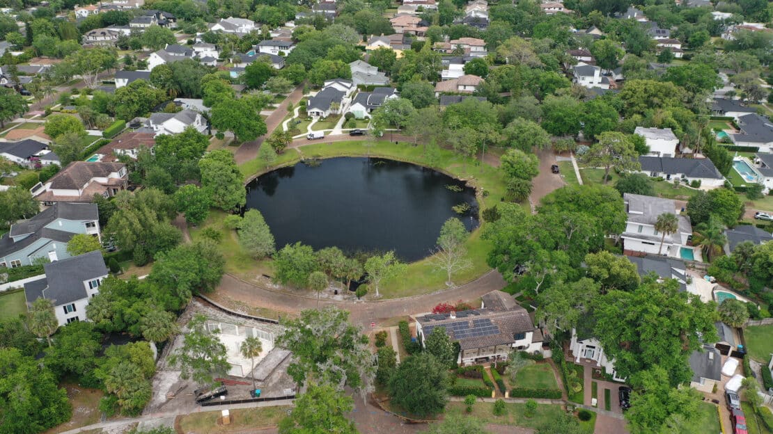 Aerial view of a residential neighborhood with a circular lake in the center, surrounded by trees and houses. Streets and greenery fill the area, creating a serene suburban landscape with scenic water views.