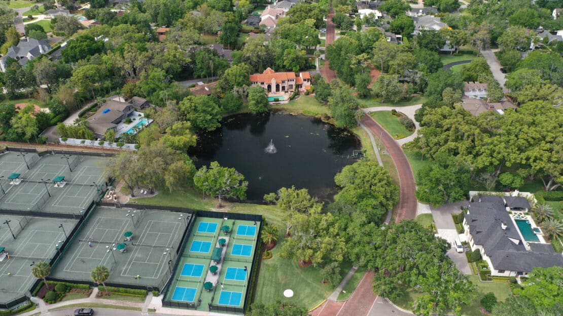 Aerial view of a suburban neighborhood featuring a small lake with a fountain, surrounded by trees, houses, tennis and pickleball courts, and winding roads.