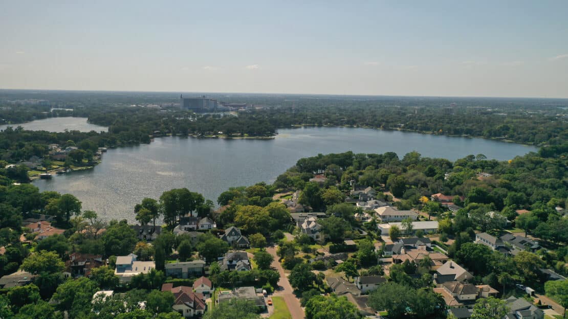 Aerial view of a suburban neighborhood with tree-lined streets near expansive lakes, surrounded by dense greenery under a clear sky.