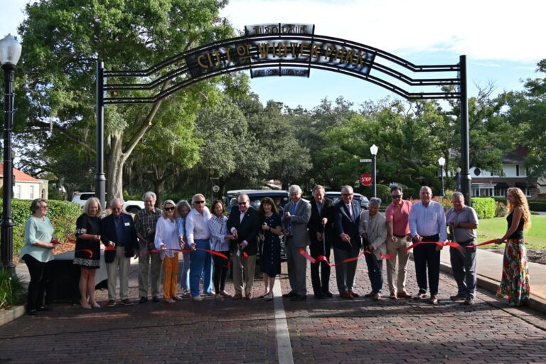 a line of people standing behind a ribbon where they are cutting it for a dedication in front of the City of Winter Park gateway sign.