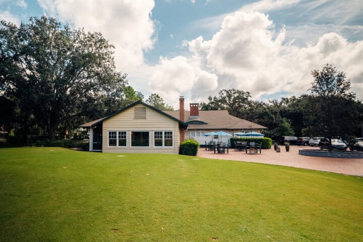Modern event venue in Winter Park featuring outdoor seating, lush green lawns, large oak trees, and a charming building with patio tables and umbrellas, ideal for community gatherings and celebrations.