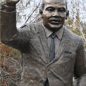 1. Bronze statue of a man in a suit gesturing with his right arm, set outdoors with fall foliage background, representing Winter Park, Florida.