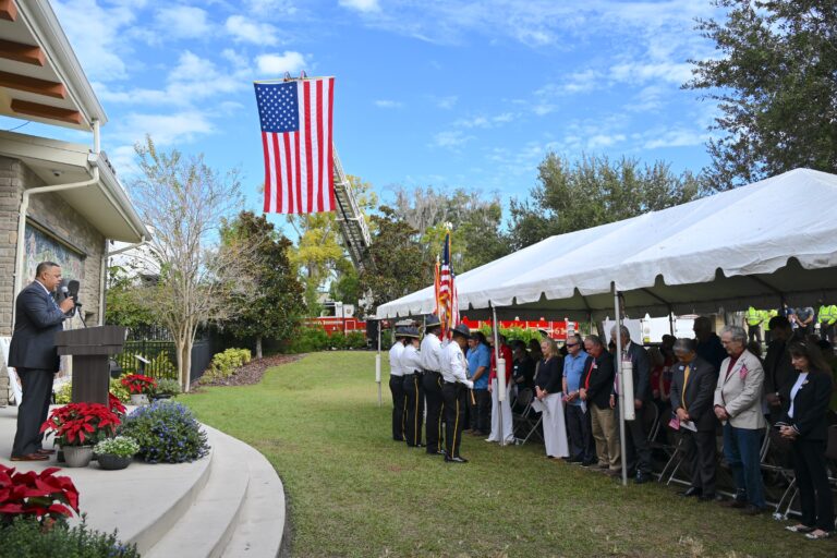 People standing under a large tent with the American flag flying from the fire-rescue truck ladder with the honor guard presenting colors while the African American pastor leads the Pledge of Allegiance at the podium